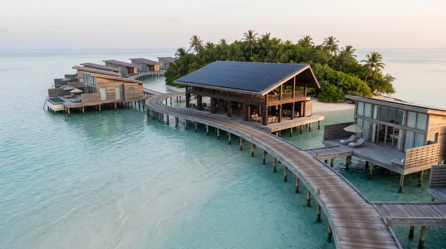 Geschwungener Steg mit Overwater-Villen aus Holz und Glas über einer hellen Lagune